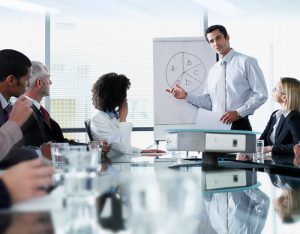 A man in a suit gives a presentation to his coworkers, displaying a poorly drawn pie chart.